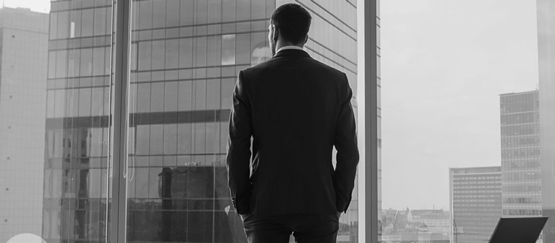 Back View of the Thoughtful Businessman wearing a Suit Standing in His Office, Hands in Pockets and Contemplating Next Big Business Deal, Looking out of the Window. Big City Business District Panoramic Window View.